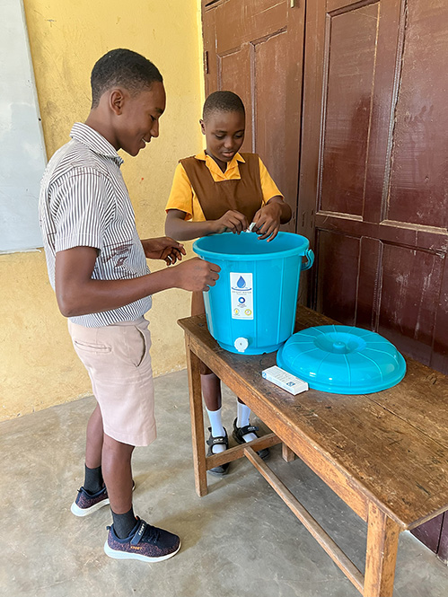 A girl and boy student treat their classroom drinking water container with a chlorine tablet.